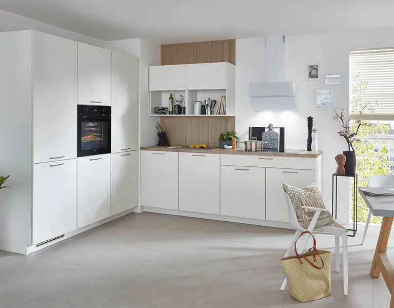 Moderne L-Küche mit weißen Fronten, Holzarbeitsplatte, integriertem Backofen und offenen Regalen, ergänzt durch dekorative Accessoires und ein großes Fenster.